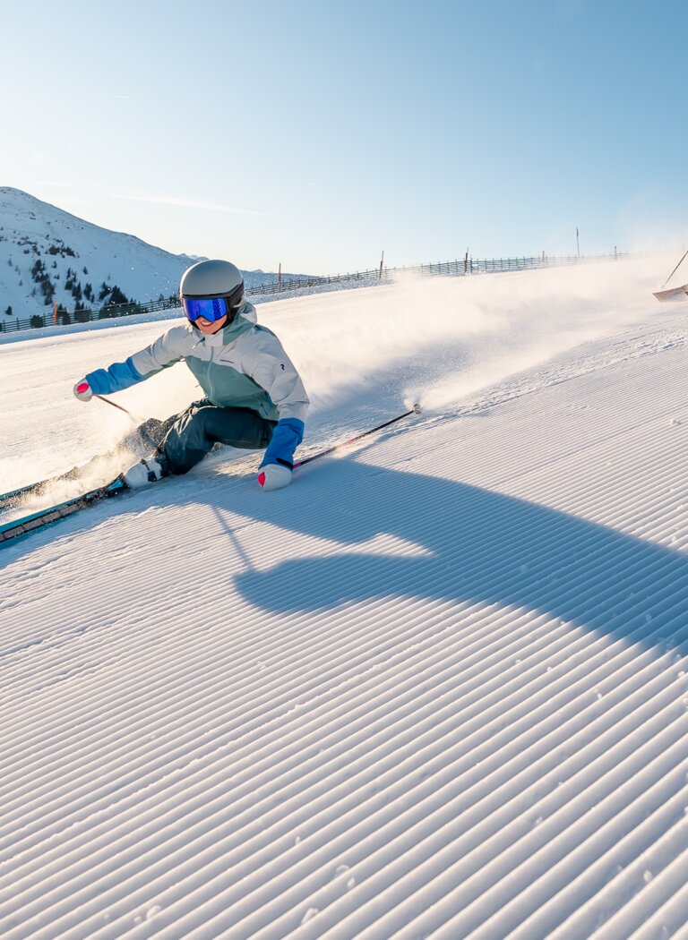 Two skiers carve turns on a freshly groomed piste at sunrise with snow-covered mountains in the background. | © Miriam Lottes