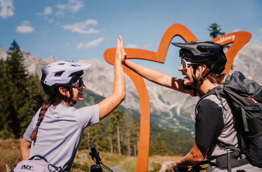 Two female mountain bikers give a high five with helmets and bikes in front of Alpine scenery | © Hochkönig Bergbahnen GmbH