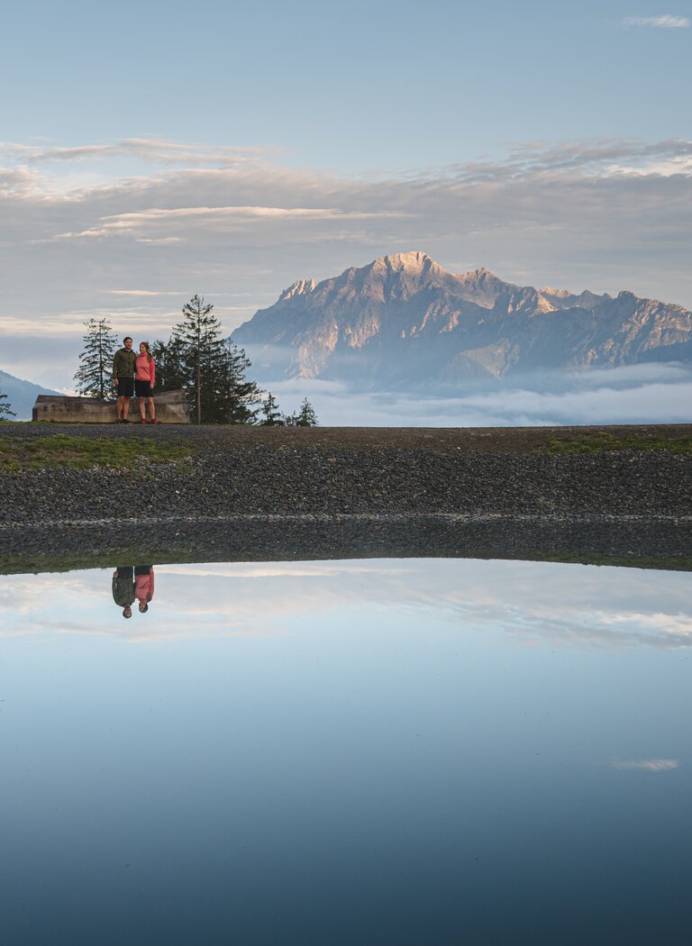 Zwei Personen stehen vor Bergpanorama mit Spiegelung im ruhigen See unter wolkigem Himmel | © Hochkönig Bergbahnen GmbH