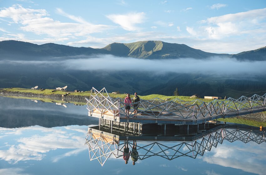 Two people stand on a pier by a calm mountain lake with reflections, mountains and low clouds | © Hochkönig Bergbahnen GmbH