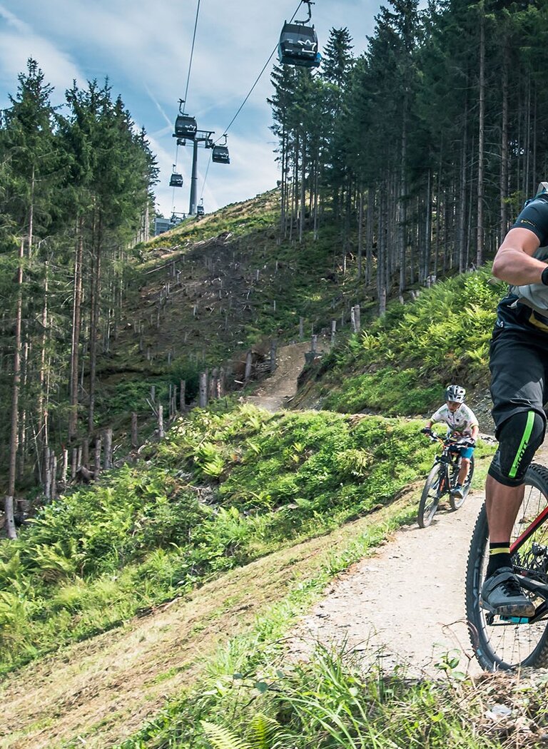 Two mountain bikers ride a flow trail through forested terrain with a gondola lift in the background. | © Hochkönig Bergbahnen GmbH
