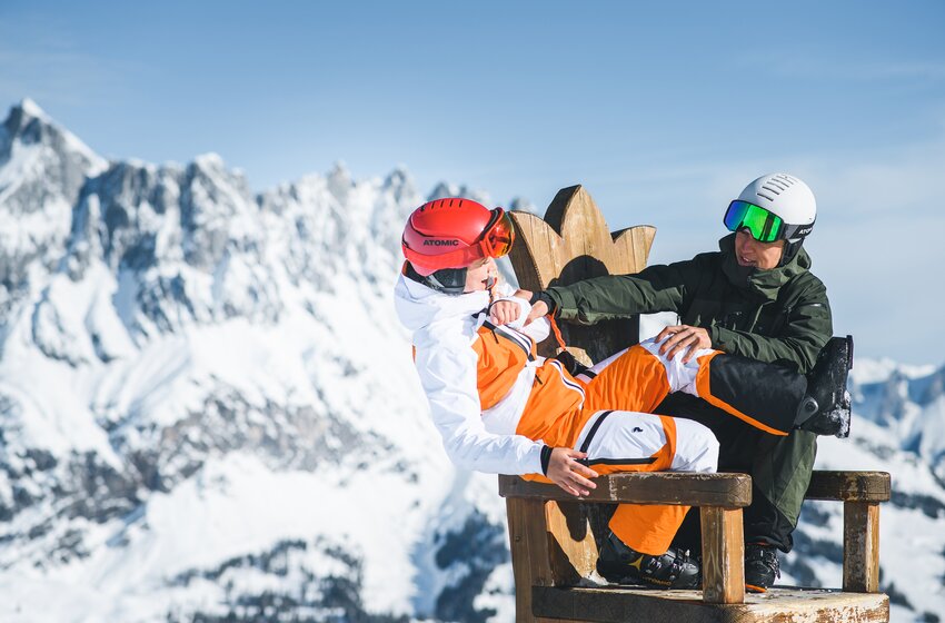 Two skiers joking on a wooden bench with mountain panorama in the background at Hochkönig. | © Hochkönig Bergbahnen GmbH