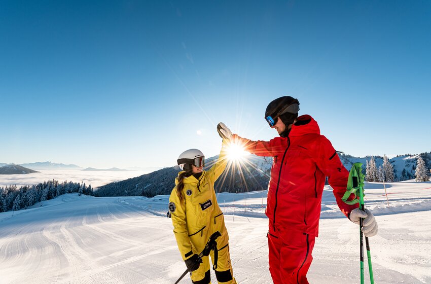 Two skiers high-five at sunrise on a freshly groomed slope in the Hochkönig ski area. | © Hochkönig Bergbahnen GmbH